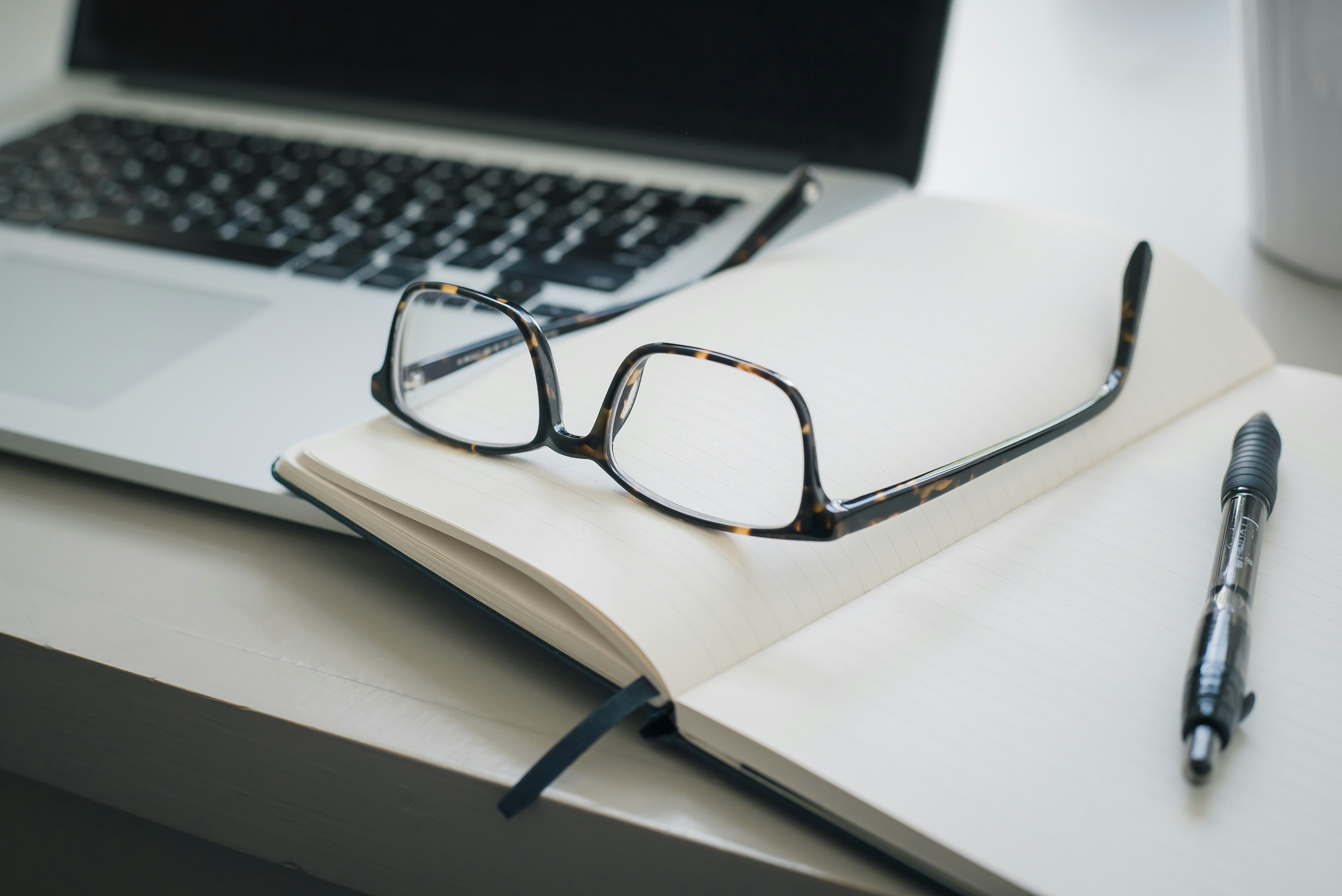 A laptop and notebook with a pen and reading glasses